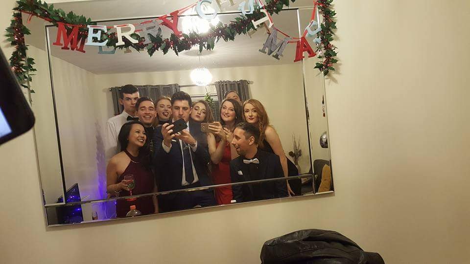 A group pose in front of a mirror to take a selfie. There is tinsel draped artfully over the top of the mirror as well as a cute felt banner that reads "Merry Christmas"